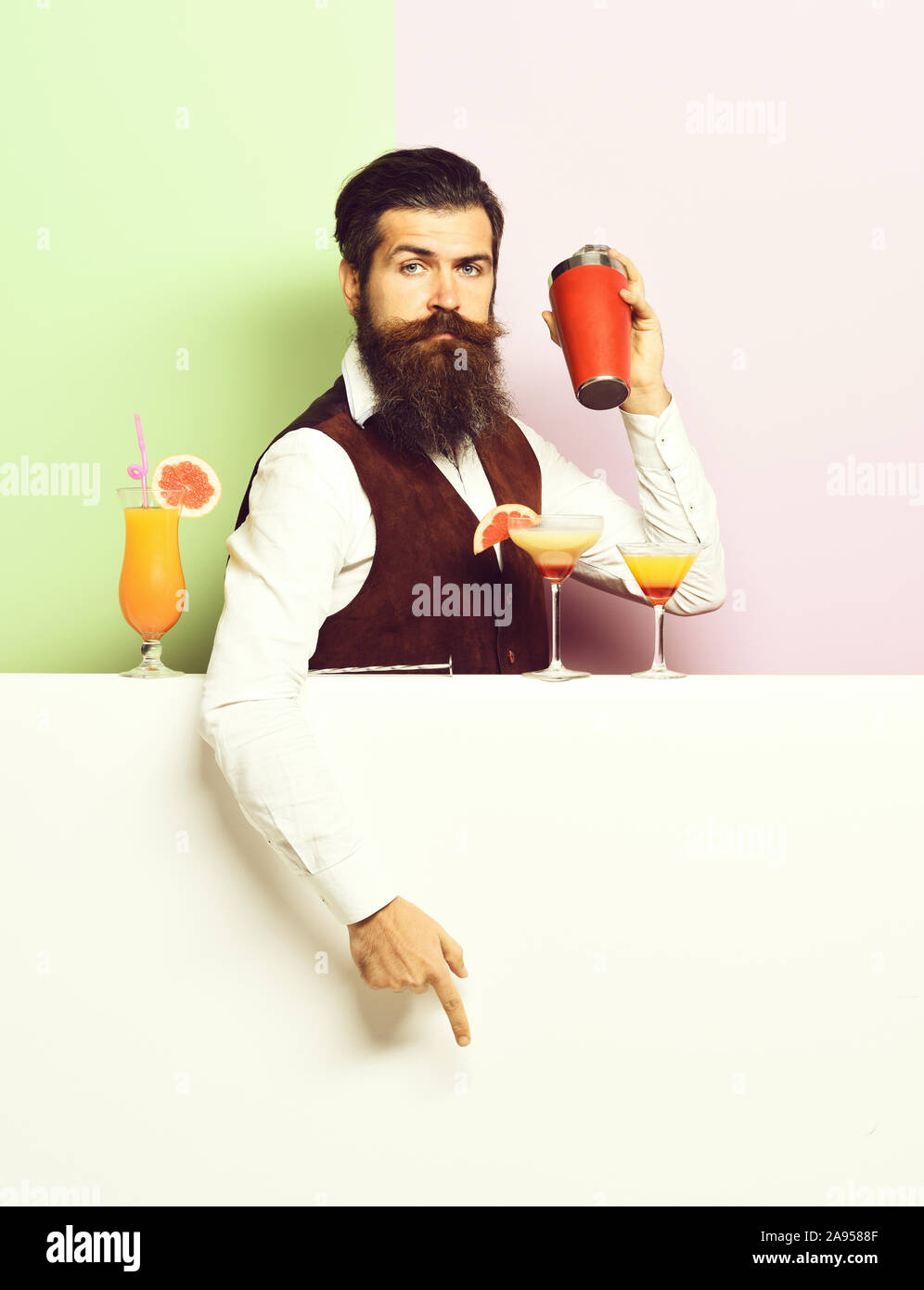 handsome bearded barman with long beard and mustache has stylish hair on serious face holding shaker and made alcoholic cocktail in vintage suede leather waistcoat. on purple green studio background Stock Photo