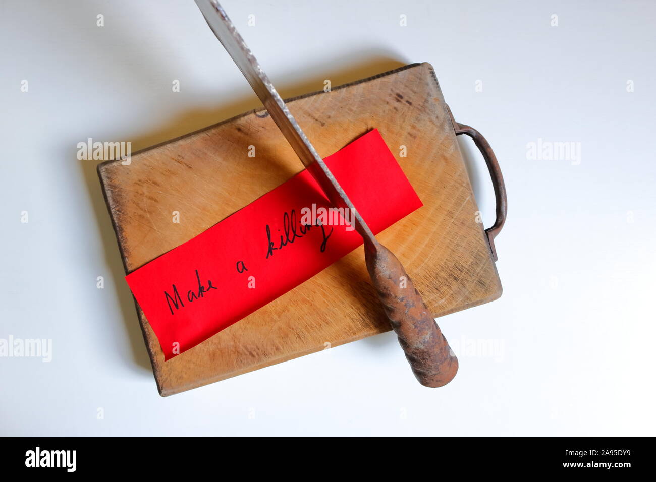 Closeup and focus on the phrase make a killing written on red paper, stuck to the cutting board with rusty chopping knife, on white wooden surface Stock Photo
