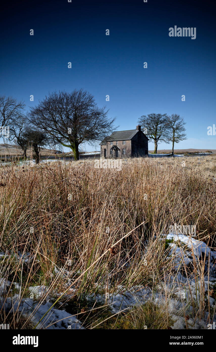 Remote solitary stone cottage surrounded by small group of trees in empty moorland landscape with light snow on ground and blue sky Stock Photo