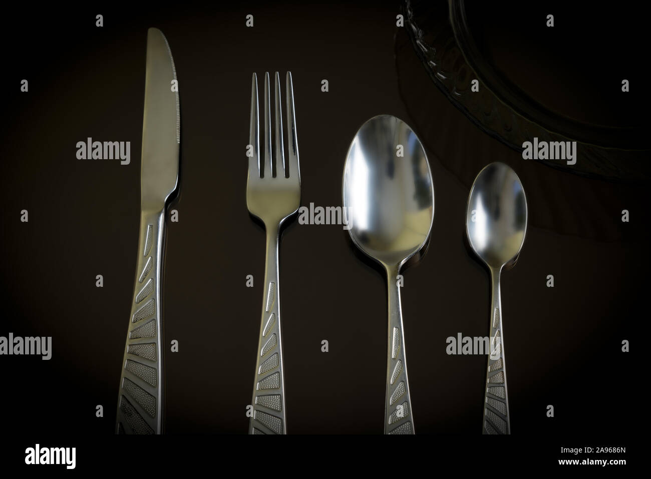Still life of the kitchen objects. Dark glass plate, silver knife, fork, spoon, teaspoon on the dark reflective backdrop. Stock Photo