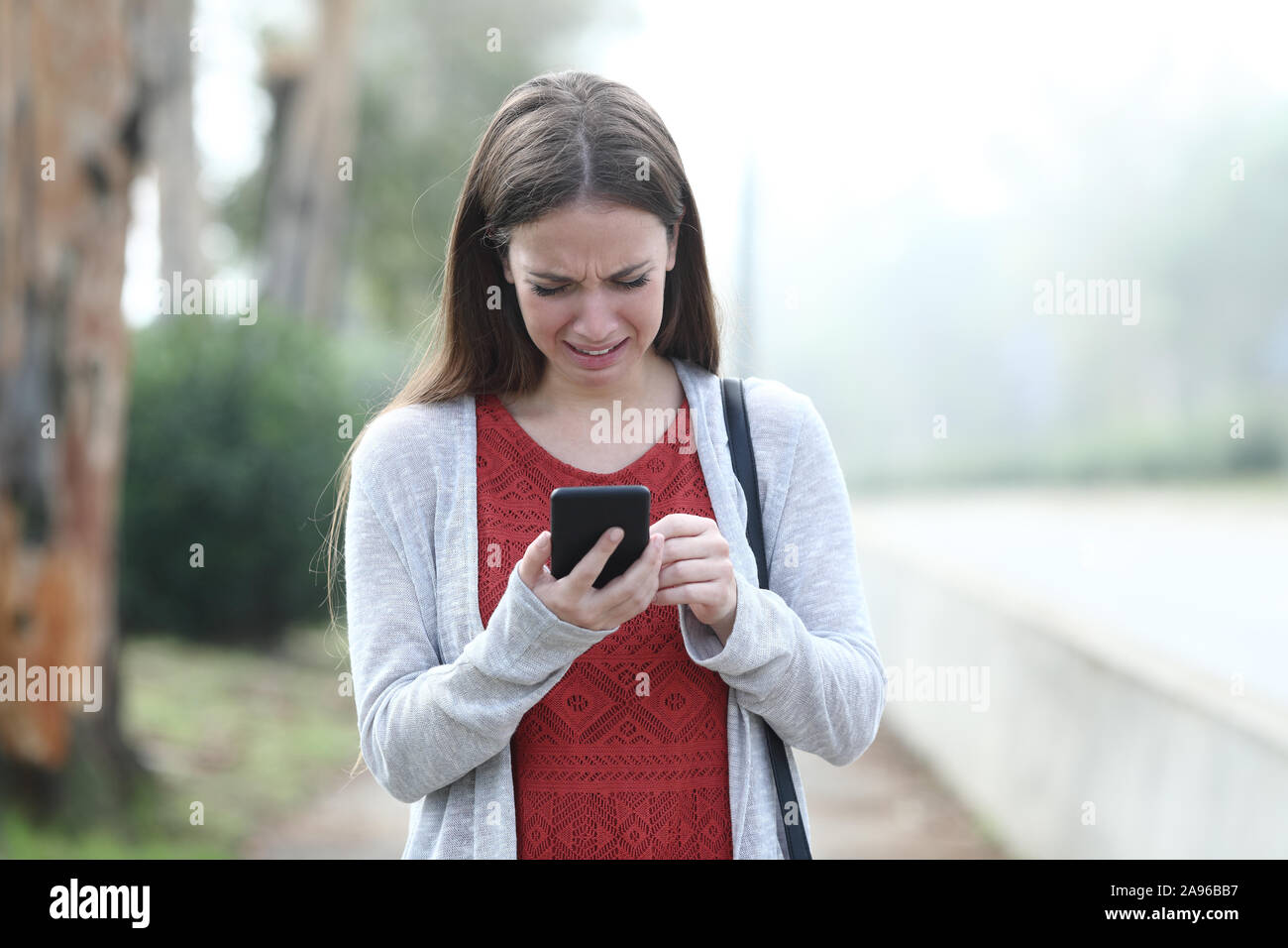 Front view portrait of an sad woman crying walking using mobile phone in a park Stock Photo