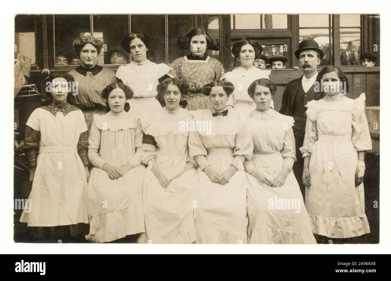 Original, clear post Edwardian era postcard group portrait of working class factory or seamstress girls working in textiles and clothing factory or workshops They are wearing white smocks/dresses, with foreman wearing a bowler hat. The photo is full of characters such as the peeking boys looking over the girls' shoulders, circa 1912 U.K. Stock Photo