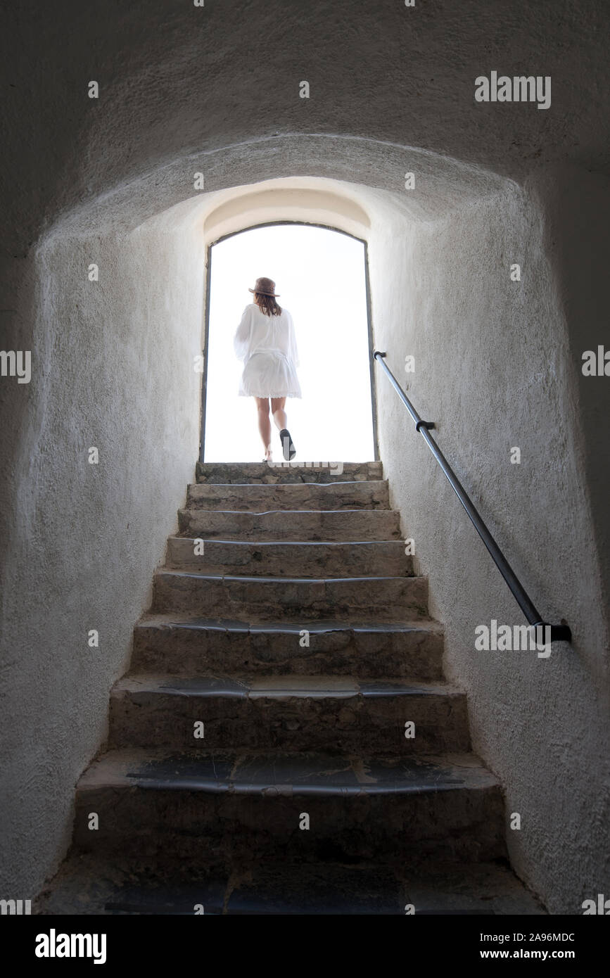 Woman walking out into daylight off a steep flight of stone stairs in an old rustic building viewed from the bottom of the steps Stock Photo
