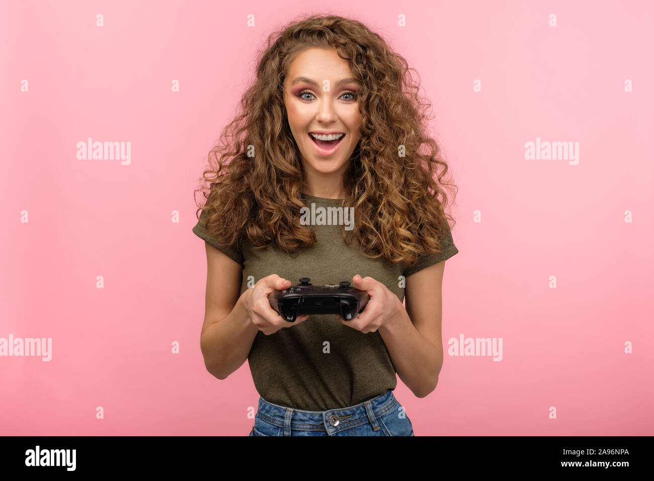 Pretty gamer girl with curly hair holding gaming controller and smile isolated on pink background Stock Photo