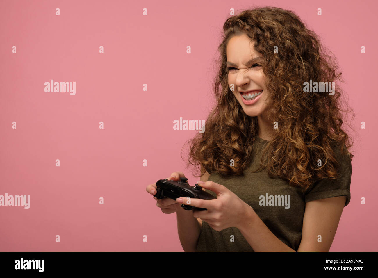 Pretty geek girl with curly hair holding gamepad and playing videogame isolated on pink background Stock Photo