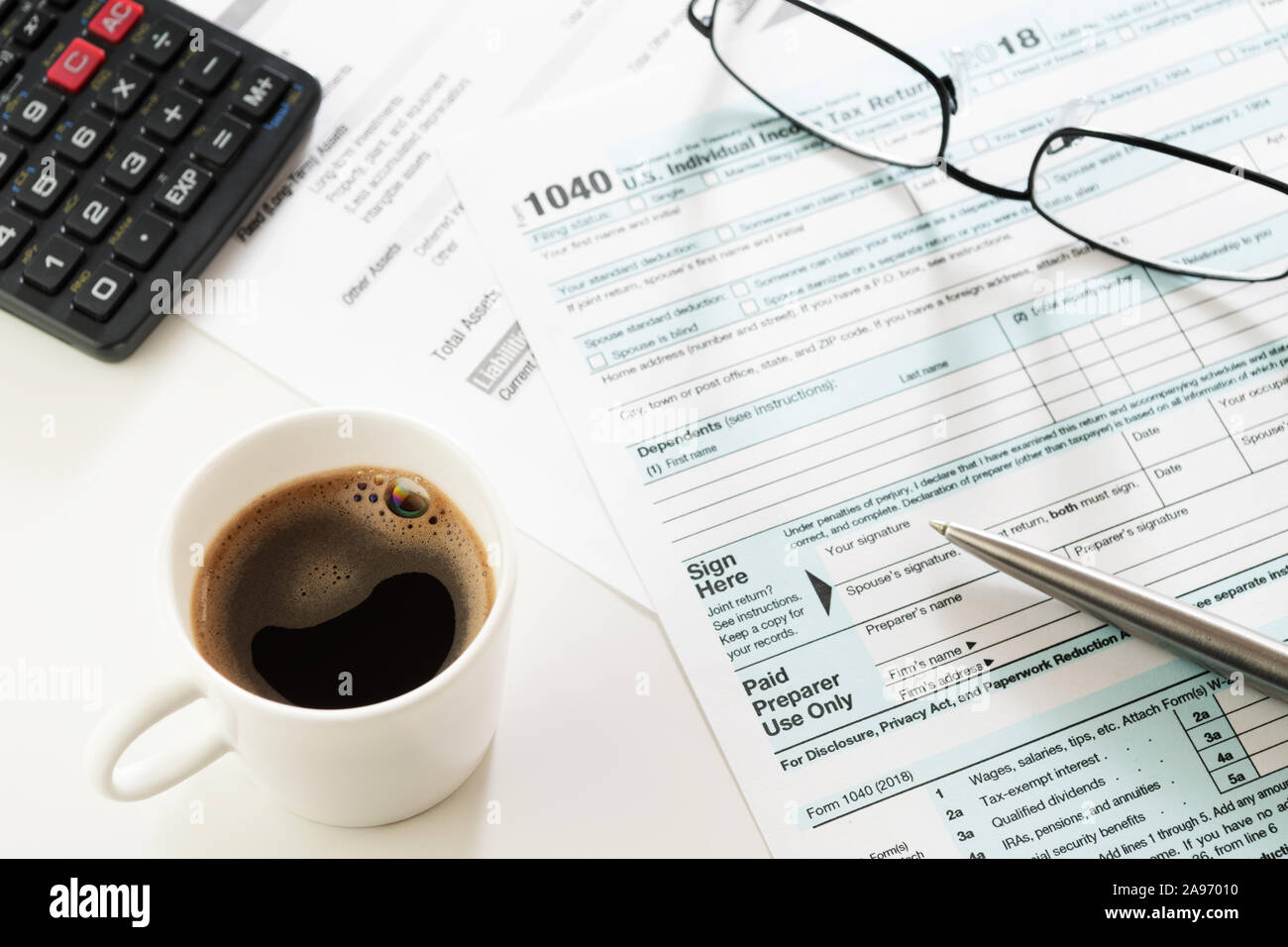 White cup of black coffee espresso on white table with visible U.S. Individual Income Tax Return form 1040, pen, calculator and eyeglasses. Taxes Stock Photo