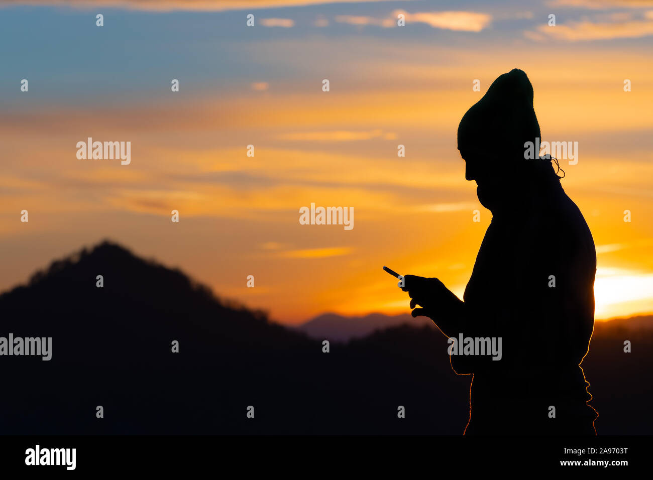 Silhouette of a male hiker with knit cap looking at his mobile phone on a beautiful sunset background. Hiking, modern technology and nature concepts. Stock Photo