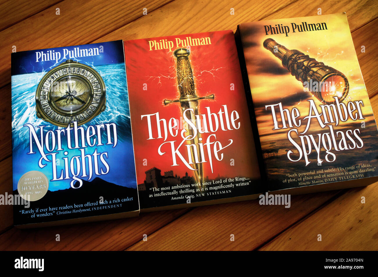 Philip Pullman His Dark Materials Trilogy. Closeup of Northern Lights, The Subtle Knife and The Amber Spyglass on a wooden table. Scholastic editions Stock Photo