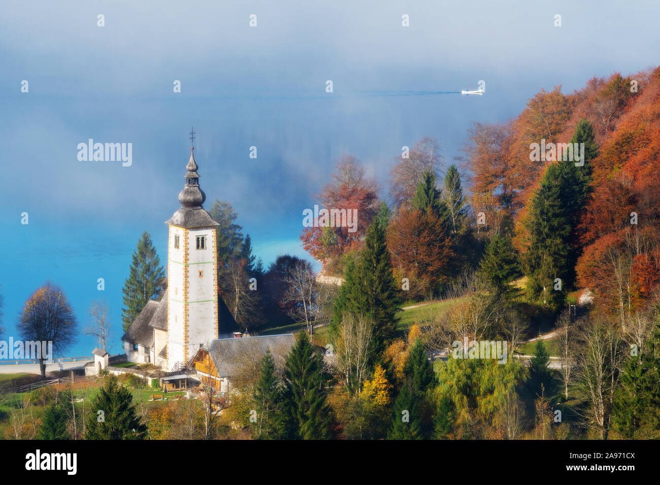 Magnificent view of catholic church of John the Baptist at lake Bohinj in autumn and canoe emerging from fog on a sunny morning in Slovenia. Travel, t Stock Photo
