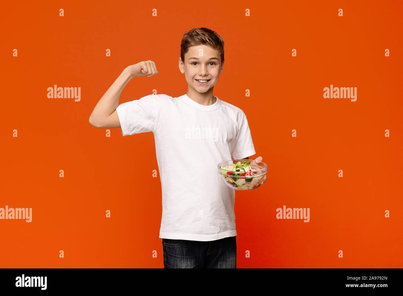 Healthy nutrition. Strong teenager with fresh vegetable salad showing his muscles, orange studio background Stock Photo