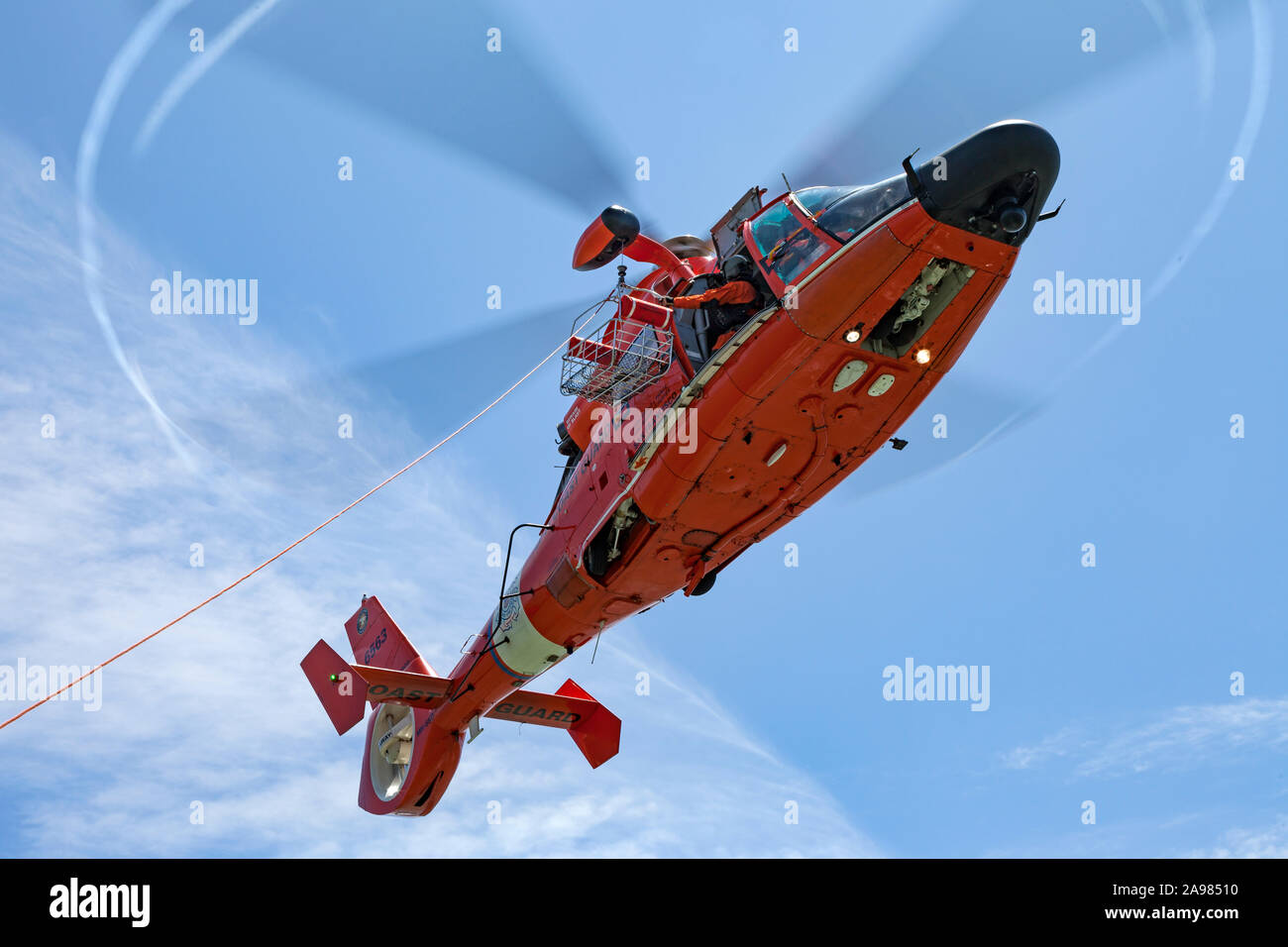 Flight Engineer looks down from the door of an USCG MH-65 Dolphin helicopter as he lowers a trail line to a vessel below. Stock Photo