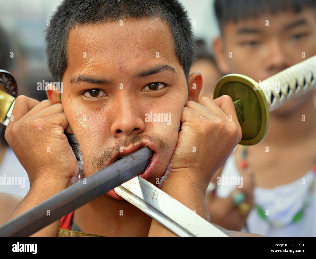 Young Thai Chinese Taoist devotee (mah song) pierces both cheeks with two long traditional swords. Stock Photo