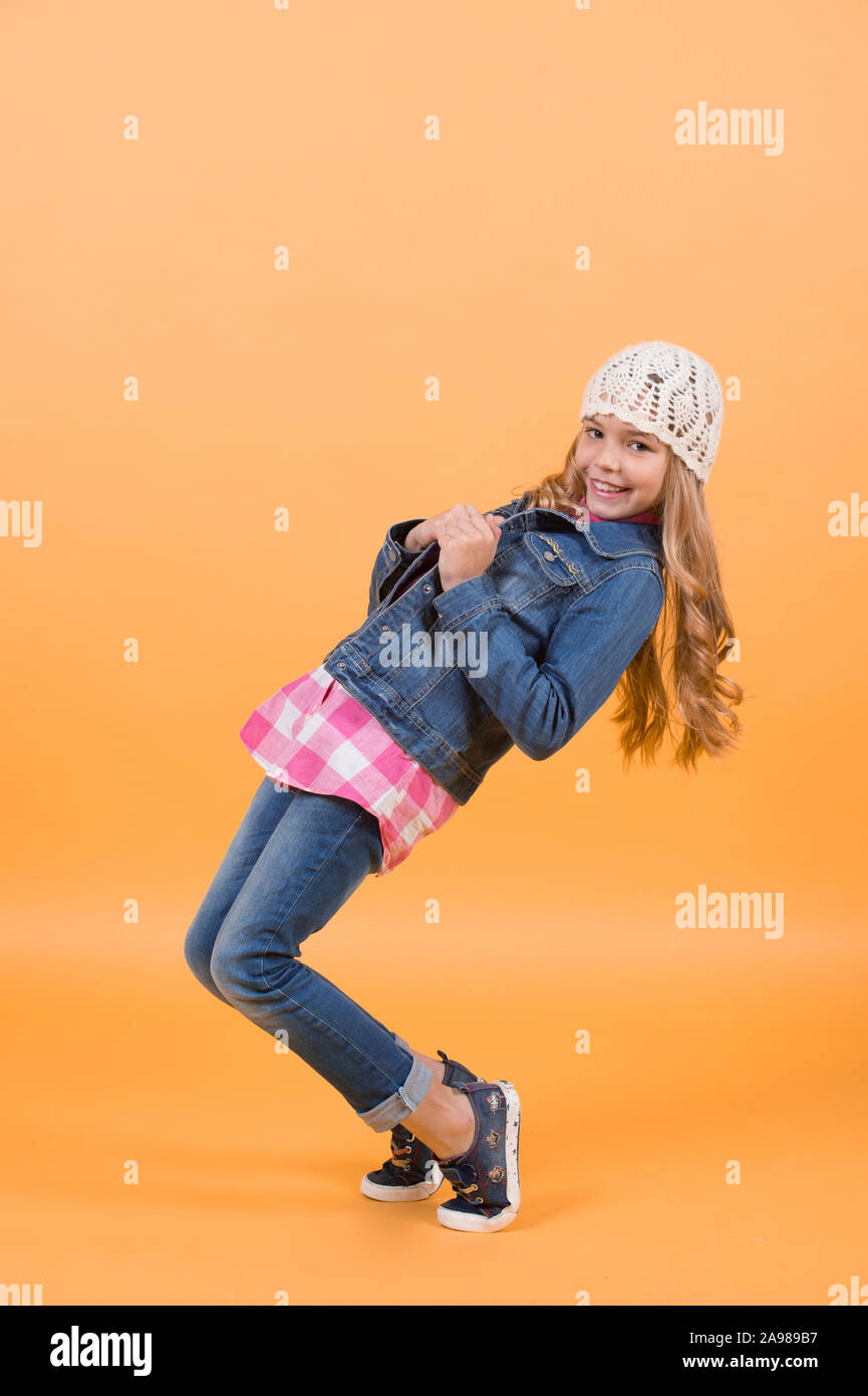 Girl model with long blond hair smile and balance tiptoe. Child in jeans suit, hat, plaid shirt on orange background. Fashion, style, trend. Beauty, look, hairstyle. Happy childhood concept Stock Photo - Girl model with long blond hair smile and balance tiptoe. Child in jeans suit, hat, plaid shirt on orange background. Fashion, style, trend. Beauty, look, hairstyle. Happy childhood concept Stock Photo -