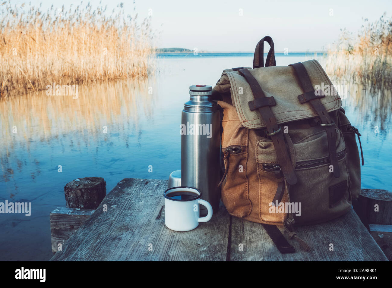 Enameled mug of coffee or tea, backpack of traveller and thermos on wooden pier on tranquil lake. Stock Photo