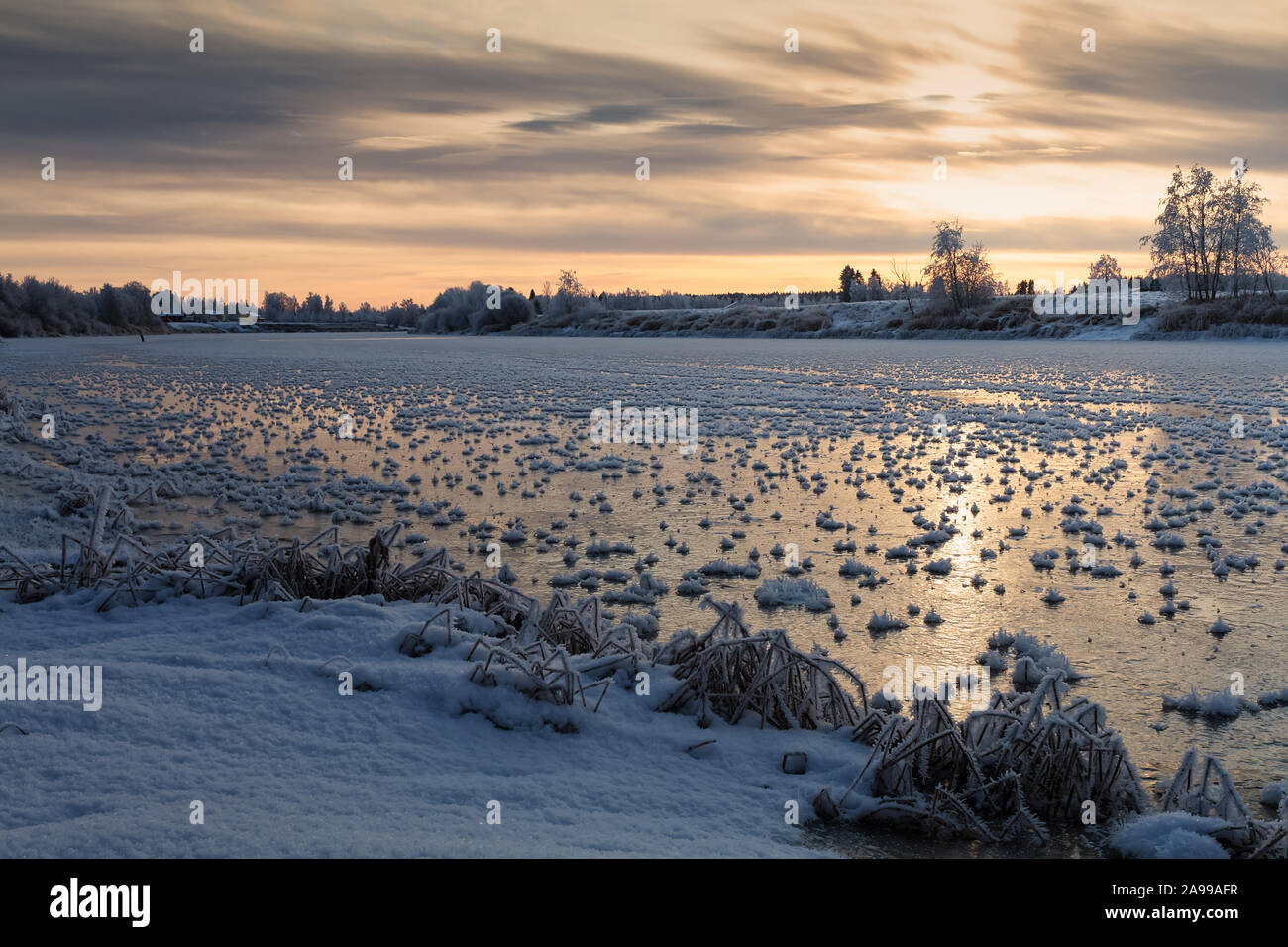 The winter sun rises over the freezing river at the Northern Finland. The river surface is covered with spiked frost formations. Stock Photo