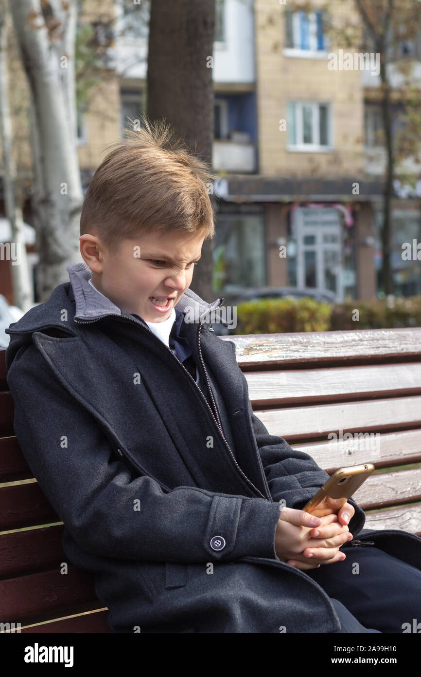 A boy with an irritated expression in a business suit sits on a bench with a smartphone.He does not like what he saw on the phone screen, he is angry Stock Photo
