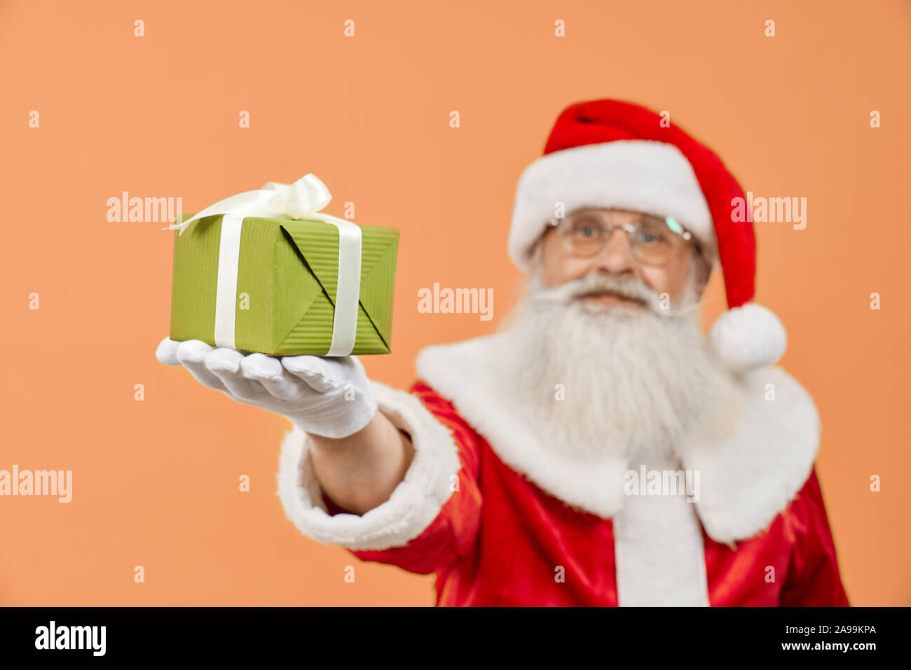 Close up of small green gift box in Santa Claus hand in white gloves and  traditional red costume on orange background. Aged mature man in eyeglasses  with real grey beard holding christmas