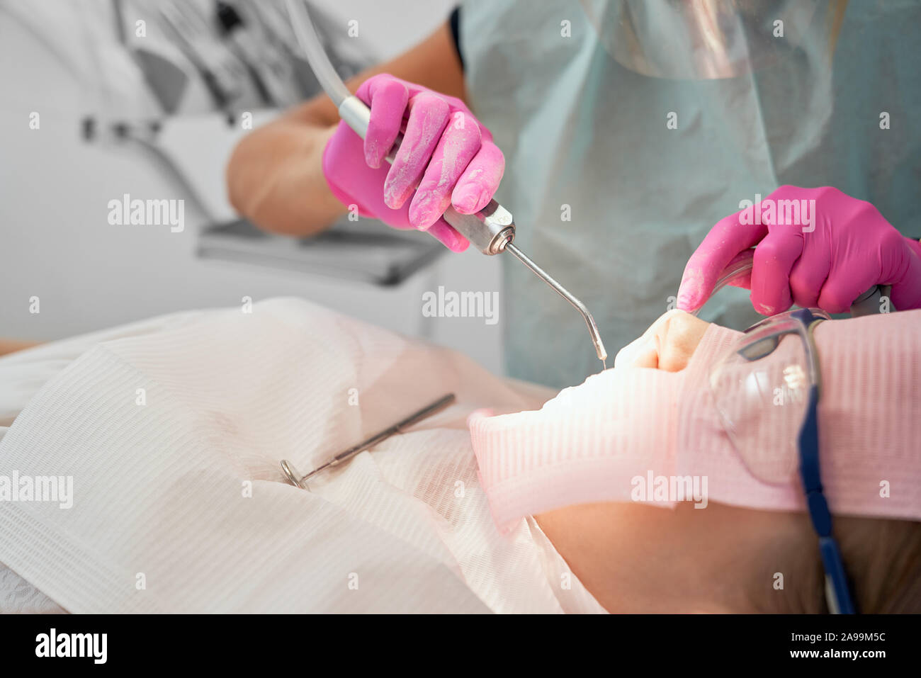 Patient of dental clinic sitting in dental chair with white bib and protective glasses. Dentist in rubber gloves holding tool, fitting teeth. Doctor doing procedure with dental curing UV light. Stock Photo