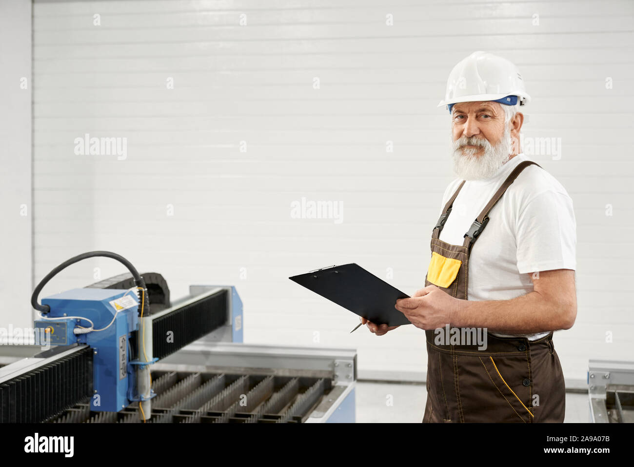 Side view of elder engineer looking at camera and posing while keeping folder and working with cnc. Professional mechanic controlling process of plasma cutting and writing data. Concept of factory. Stock Photo