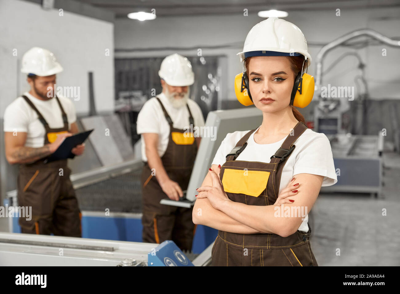 Beautiful female engineer looking at camera and posing during working day on metal factory. Young woman in helmet an uniform working with men and manufacturing products. Concept of metalwork. Stock Photo