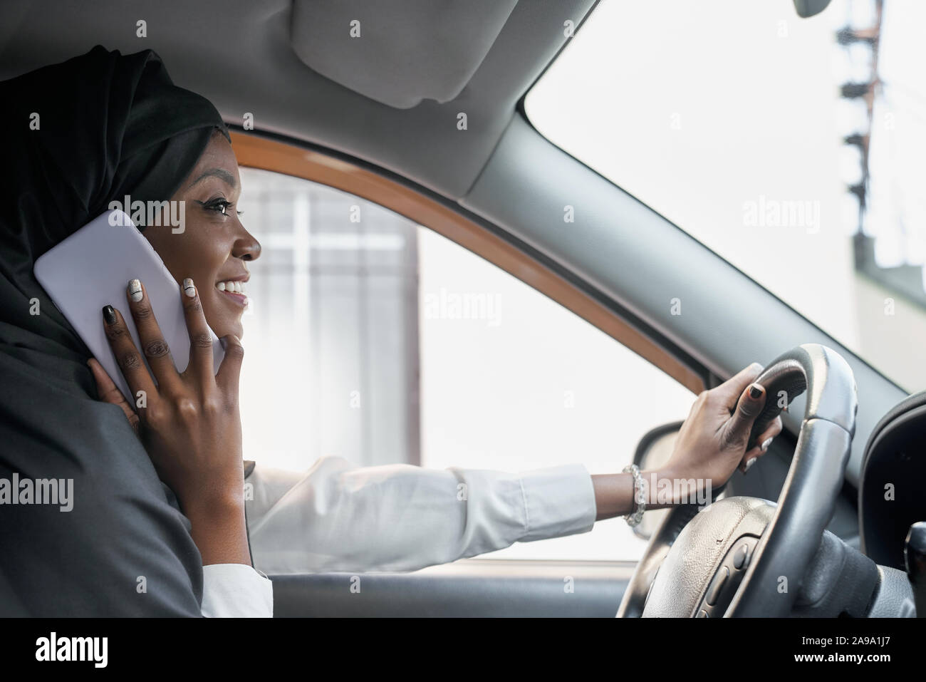 View from side of beautiful woman wearing black hijab and talking by phone while driving automobile. Religious lady keeping wheel, laughing and enjoying comfortable car. Concept of transport. Stock Photo