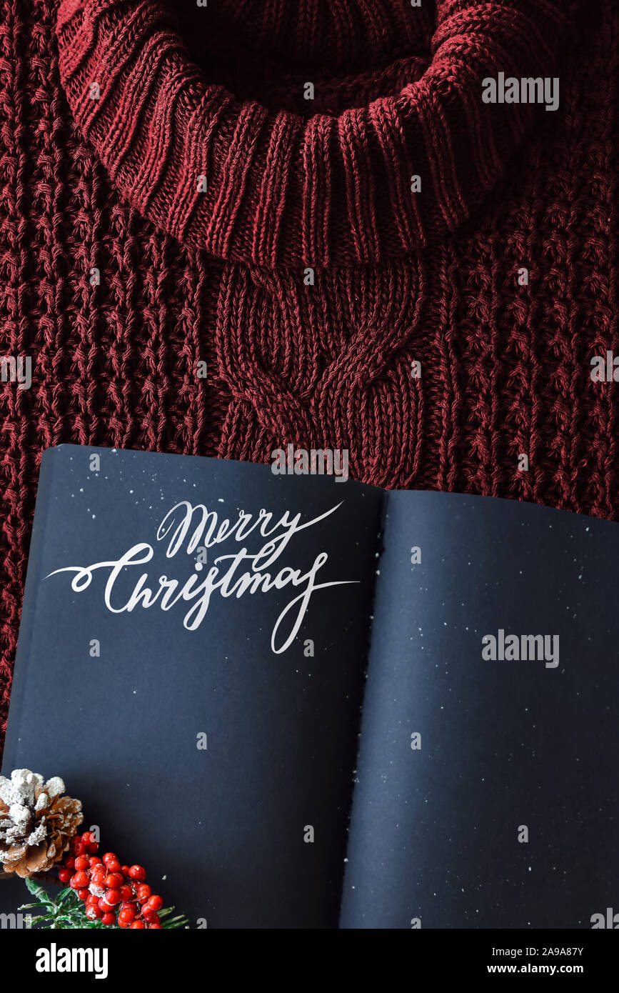 Knitted texture background of a winter red sweater with a high neck. Black notebook copy space. Hipset style. Lettering Merry Christmas. Stock Photo