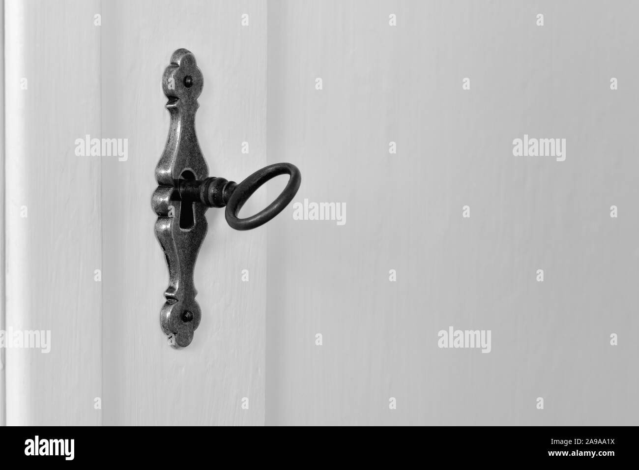 Old vintage key in the keyhole of an ornamental brass lock of a white wooden cupboard door. Monochrome, black and white, antique design, copy space. Stock Photo