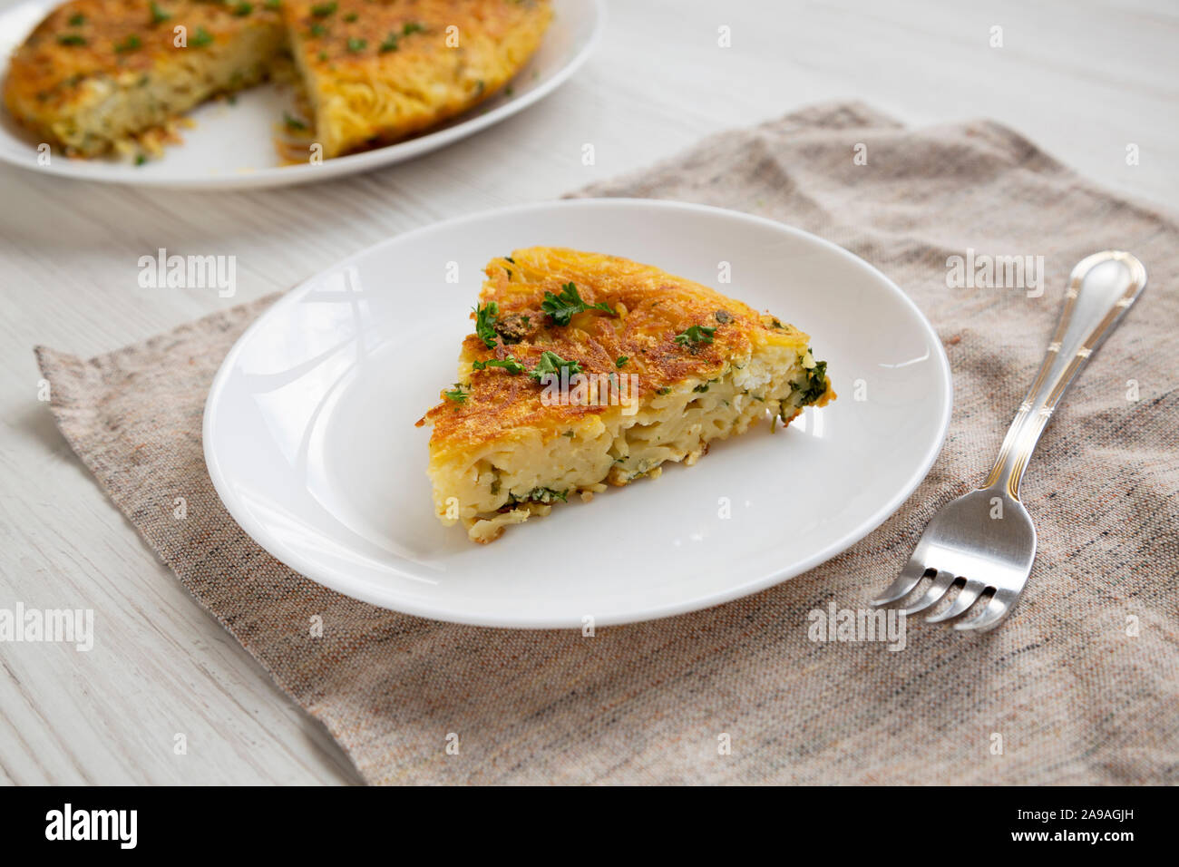 A piece of spaghetti omelette on a white plate on a white wooden surface, side view. Close-up. Stock Photo