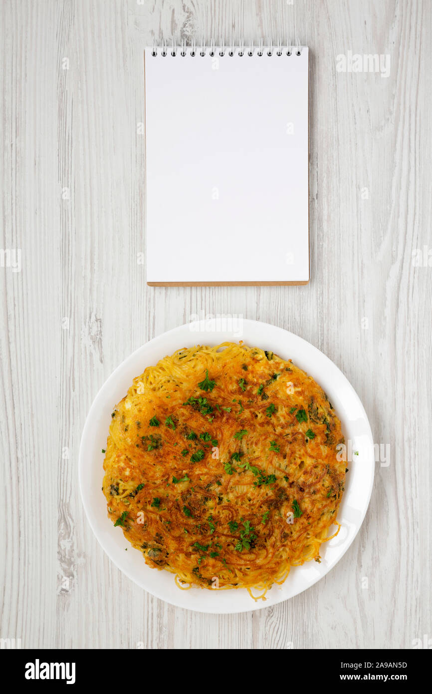 Homemade spaghetti omelette on a white plate, blank notepad over white wooden background, overhead view. Flat lay, top view, from above. Stock Photo