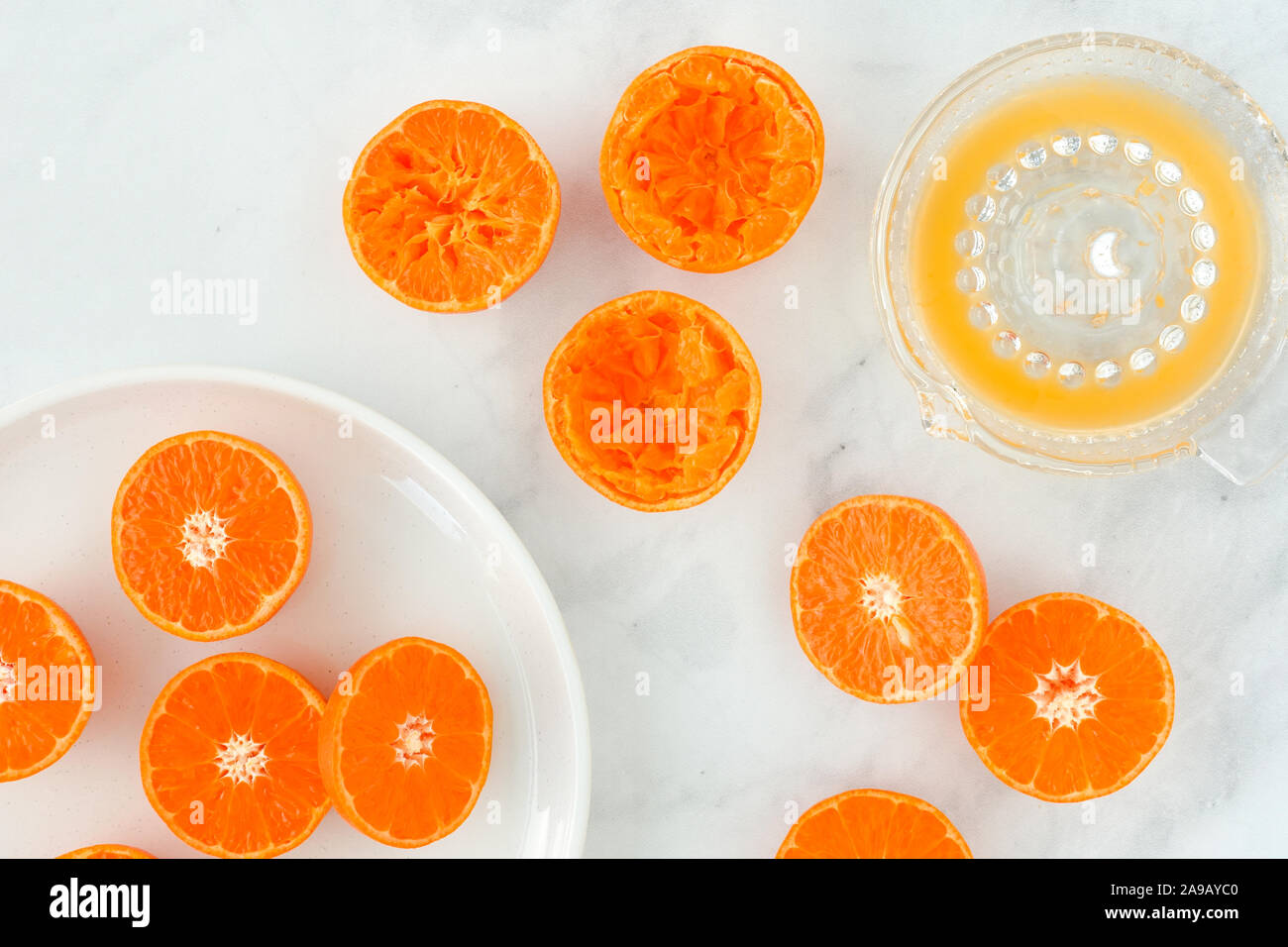 Close-up of citrus fruits, clementines, cut in half on plate and glass citrus juicer filled with juice on white marble background with copy space. Top Stock Photo