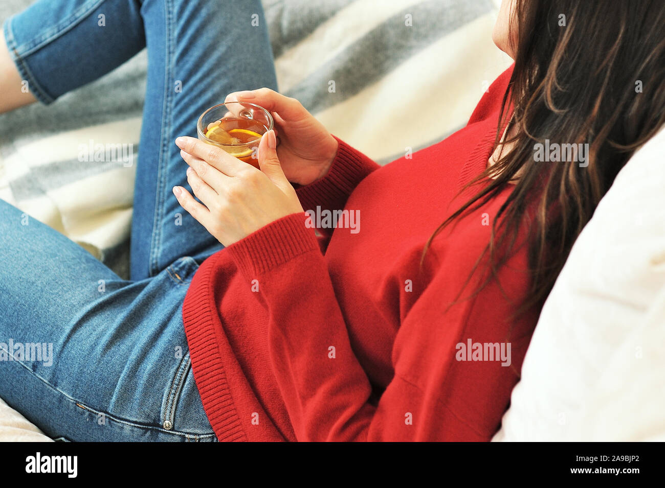 Young woman relaxing on sofa holding cup with lemon tea. Woman dreaming in living room lying on couch. Woman thinking while drinking lemon tea at home Stock Photo