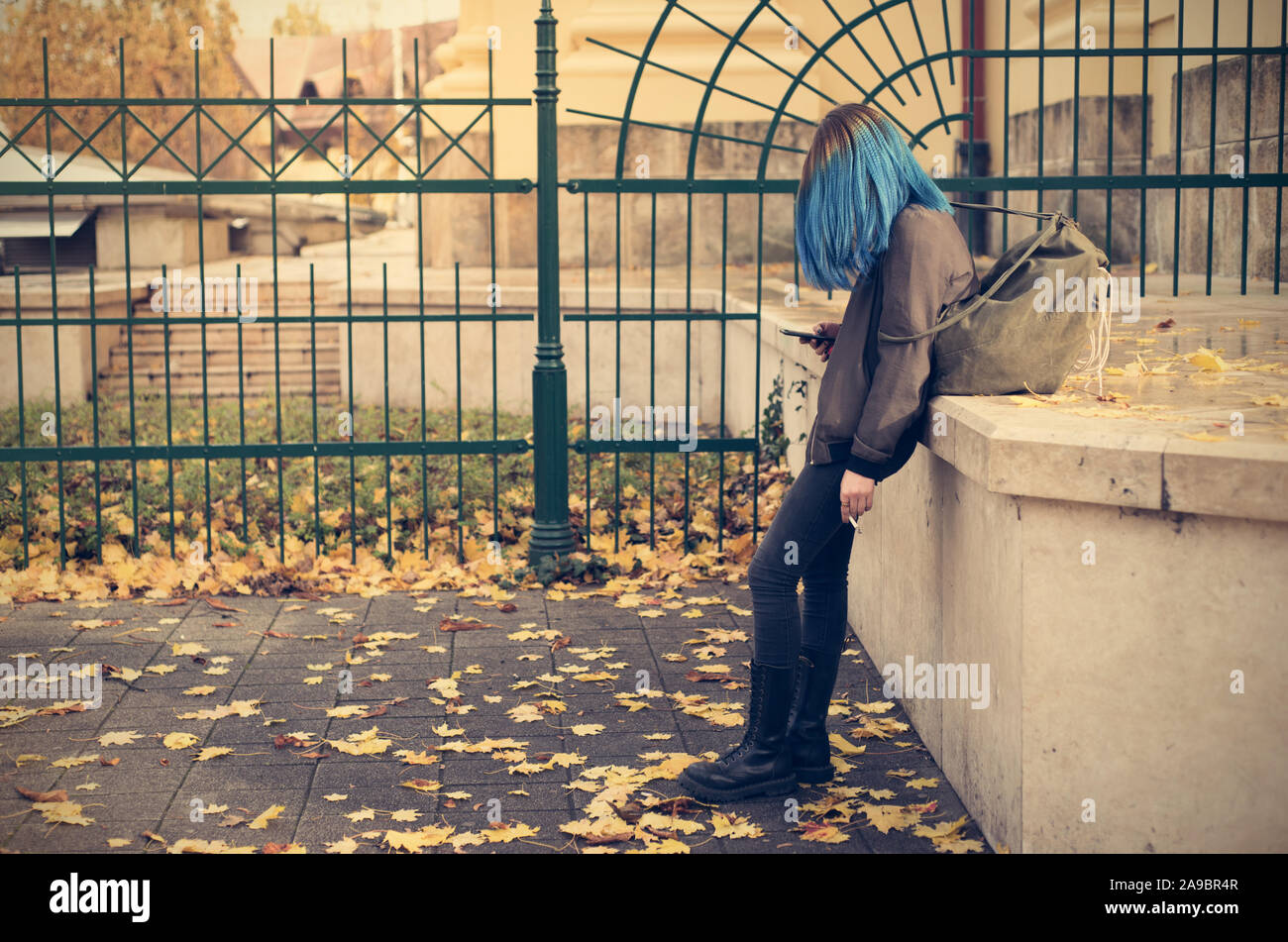 Emo girl smoking and use her phone in the street. Young student with blue colorful dyed hair Stock Photo