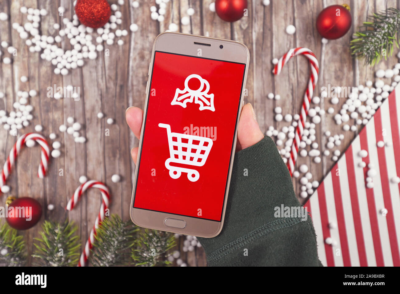 Concept for Christmas seasonal online gift shopping with hand holding cell phone with white shopping cart sign on red background in front of wooden de Stock Photo