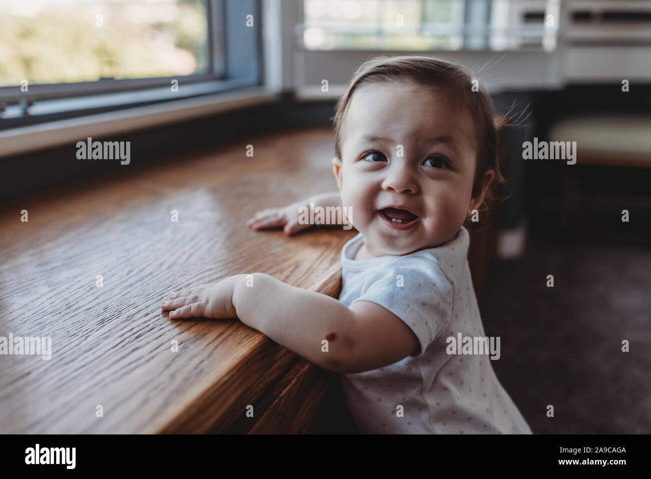 Happy baby with first teeth holding herself upright near window Stock Photo