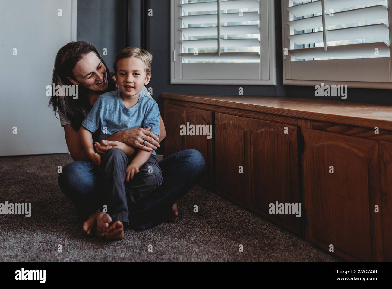 Smiling mom and son sitting on carpeted floor next to window Stock Photo
