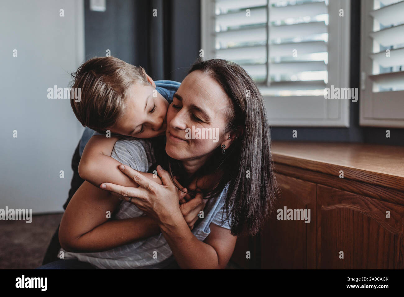 Loving hug between mid-40â€™s mom and 5 yr old son near widow shutters Stock Photo