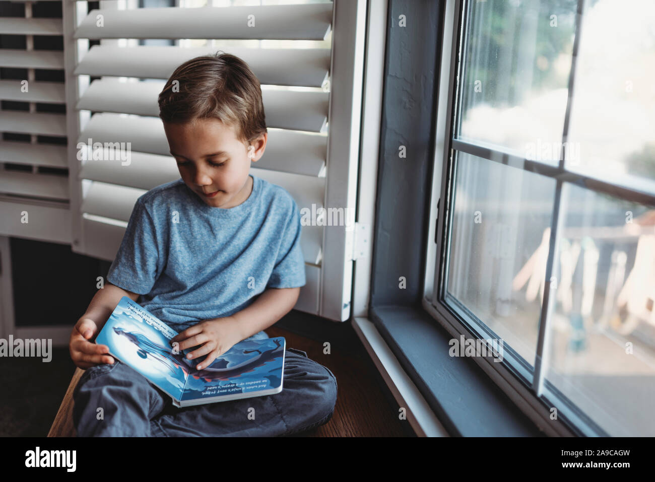 Young boy reading book by window with shutters Stock Photo