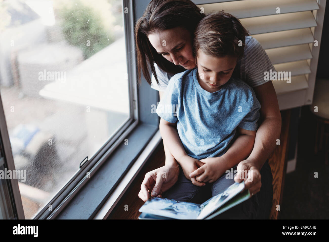 Mom reading book with son on her lap next to window Stock Photo