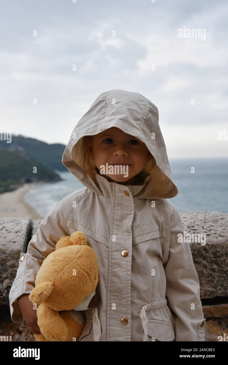 Little girl with teddy bear is wearing a raincoat jacket with hood against a stormy sky. Childhood concepts. Environmental concepts. Climate concepts. Stock Photo