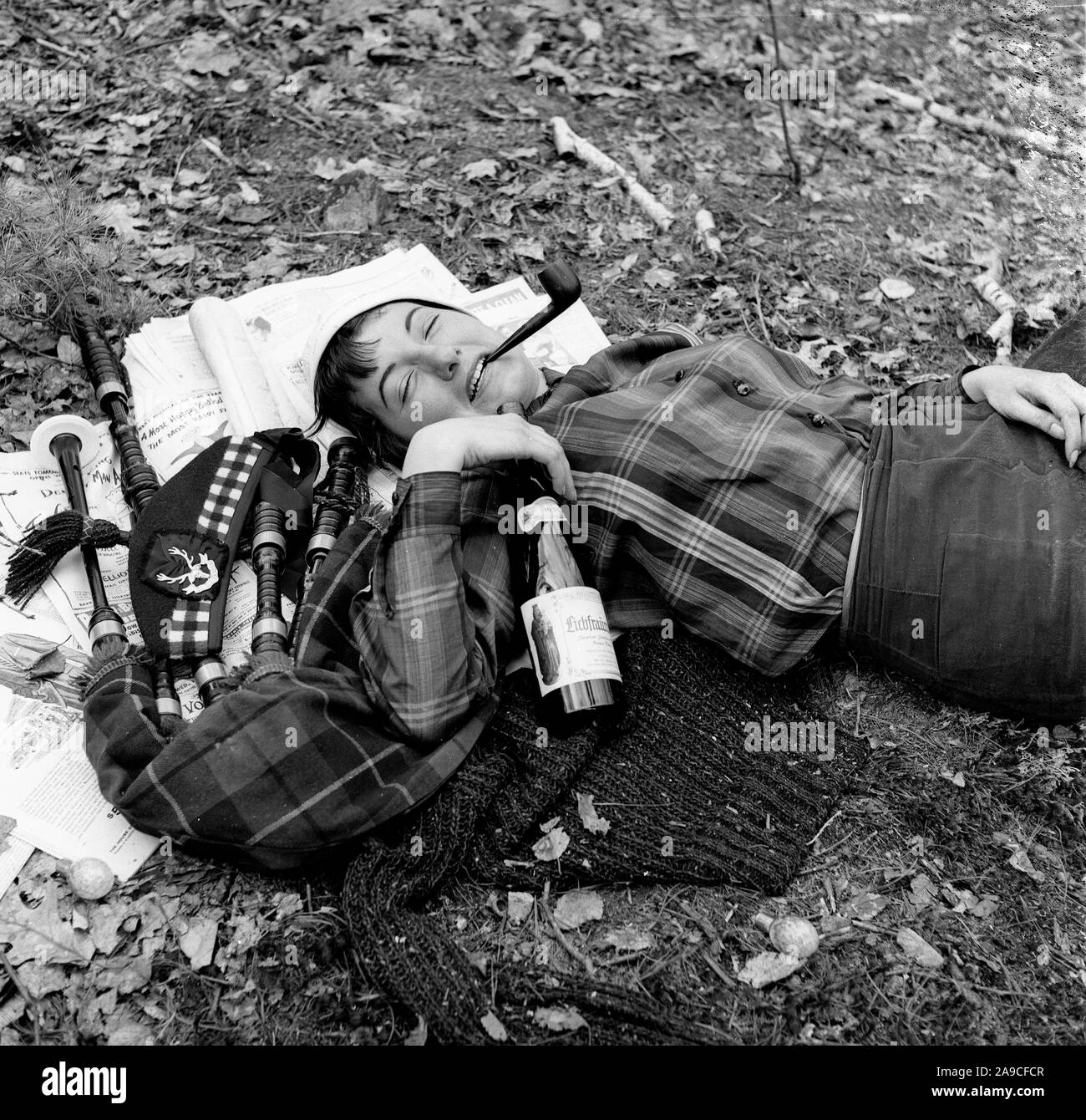 Young woman larking around with pipe in her mouth and a bottle of wine next to Gordon Highlanders cap and bagpipes USA 1956 Stock Photo