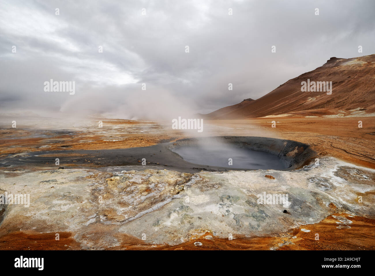 Wide volcanic landscape with a small mud crater and steam blown sideways by the wind, bright natural colours with red tones, evening light, partly clo Stock Photo
