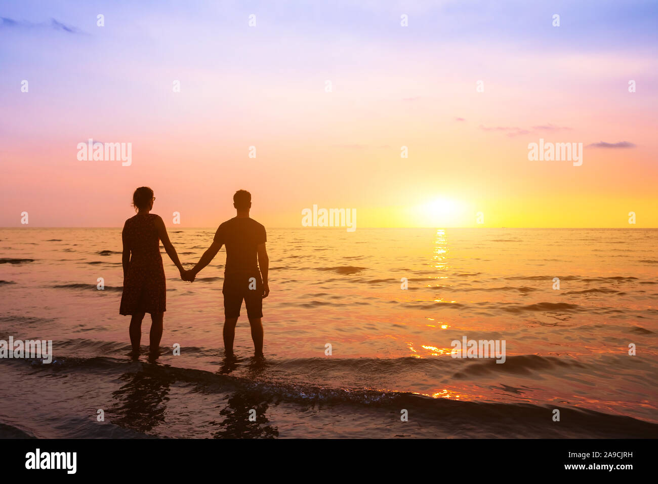 Romantic couple on the beach at sunset watching horizon, honeymoon vacation holidays at sea destination, silhouette of two lovers holding hand Stock Photo