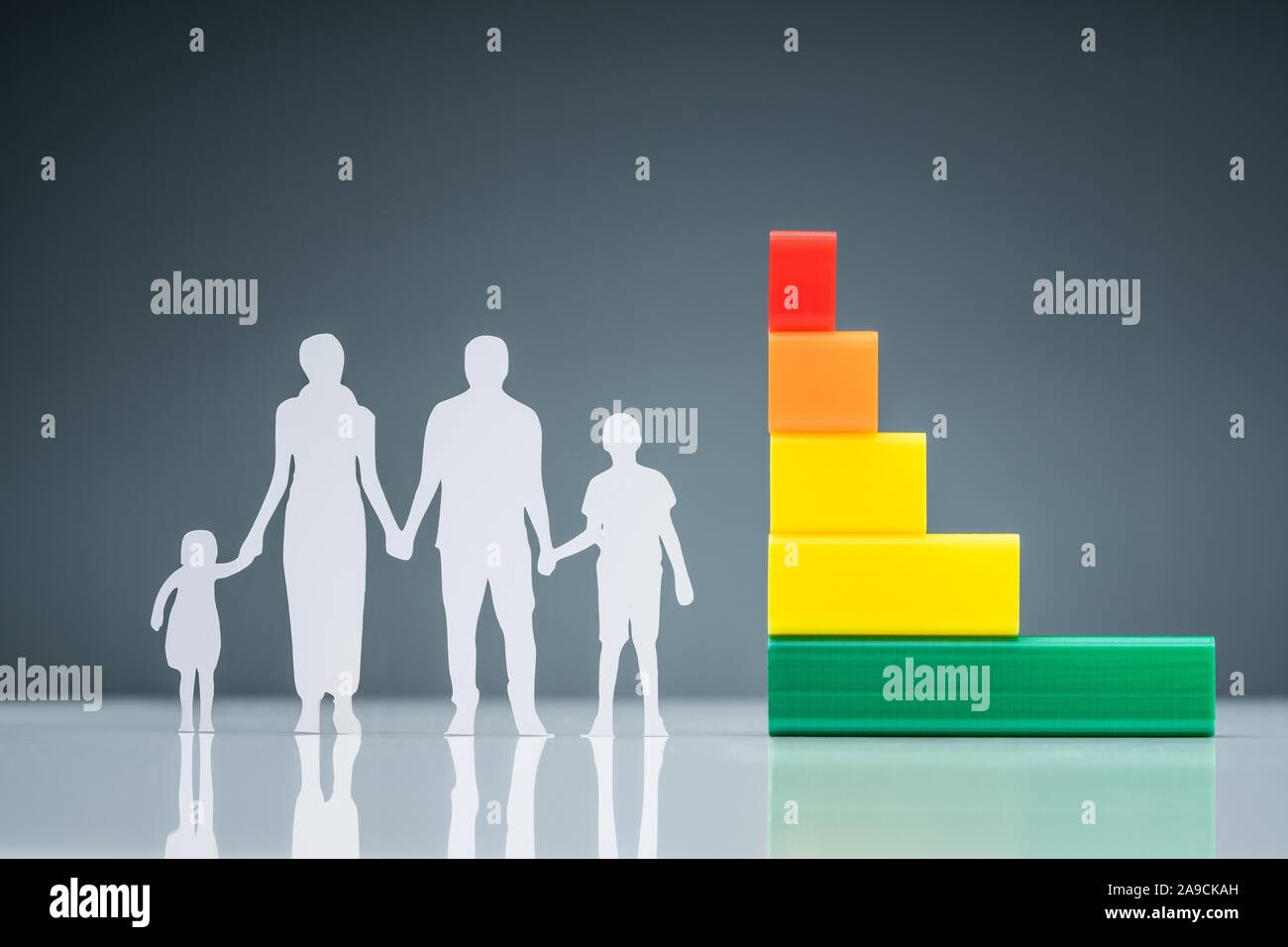 White Family Paper Cutout Near The Energy Efficiency Graph On Reflective Desk Against Gray Background Stock Photo