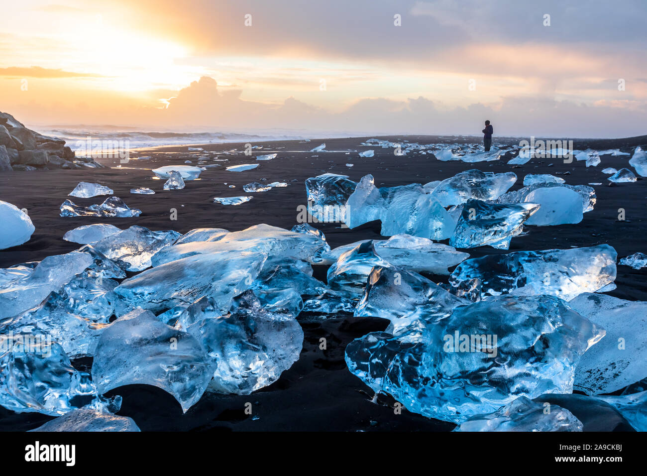 Diamond Beach in Iceland with blue icebergs melting on the black sand and ice glistening with sunrise sun light, tourist looking at beautiful arctic n Stock Photo