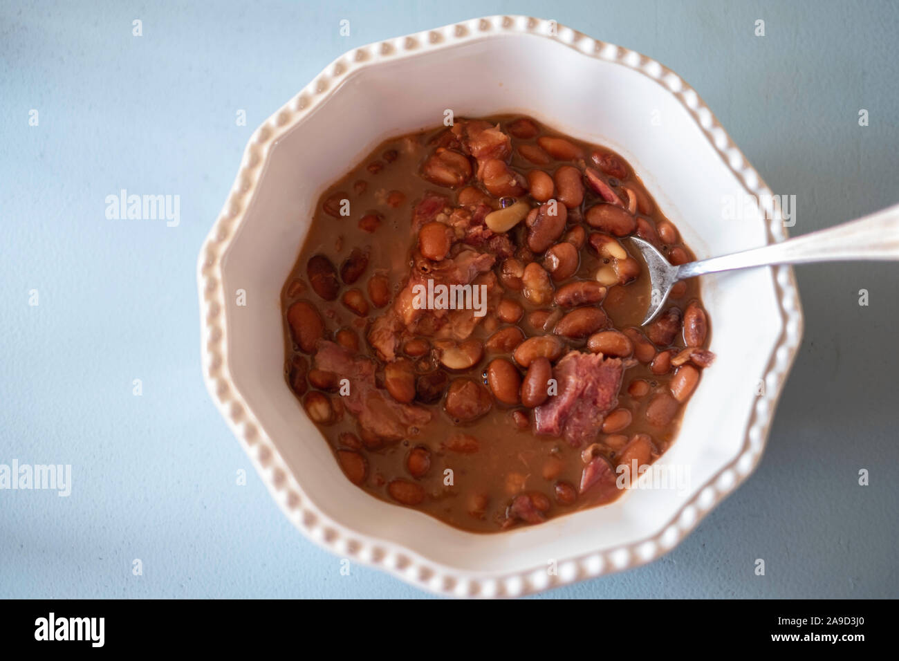 Cooked pinto beans with ham hock in a white bowl, blue background. Shot from above. Stock Photo