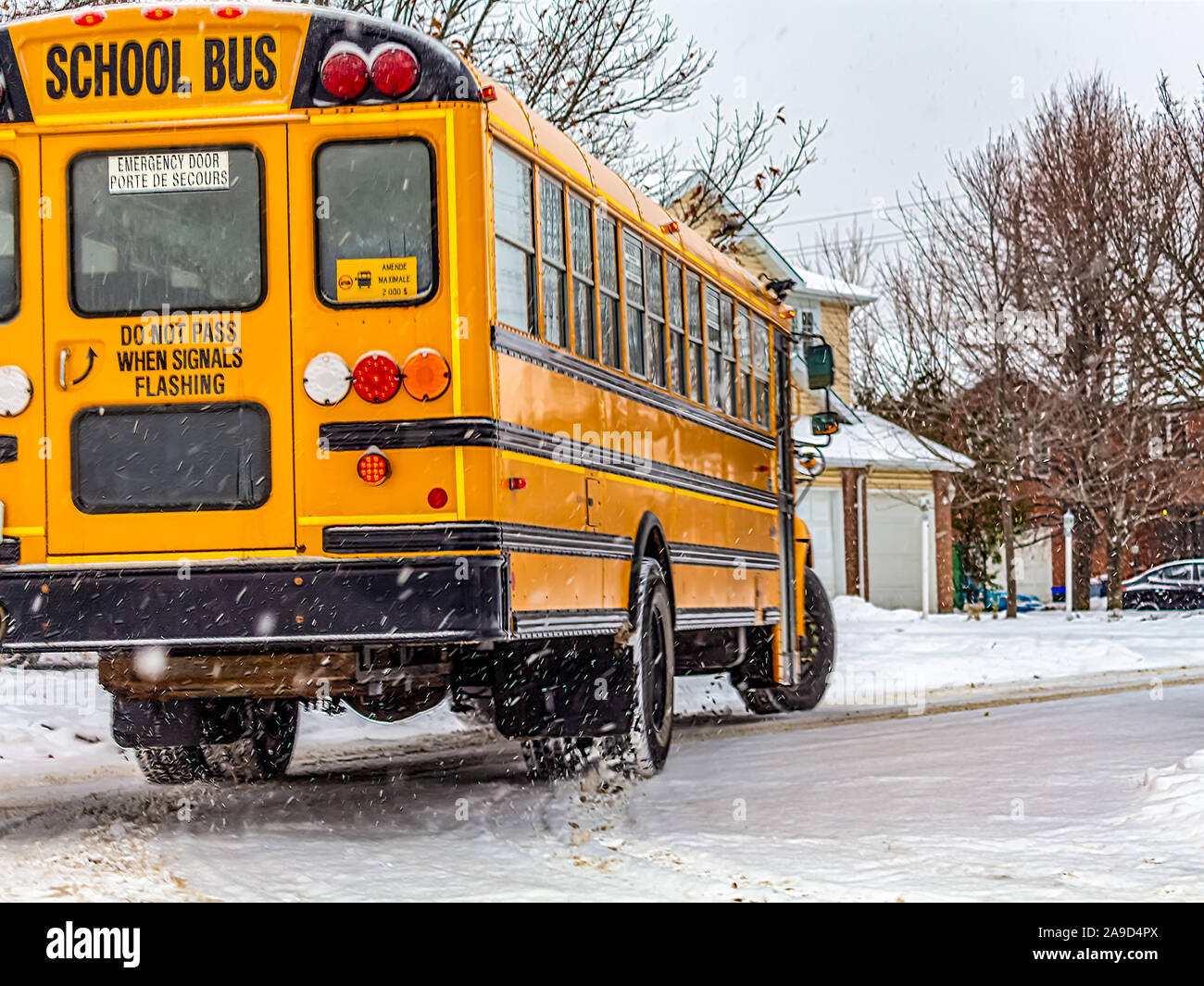 A yellow school bus turns a corner on a suburban street in the early winter. Snow has recently lined the street with white powder. Stock Photo