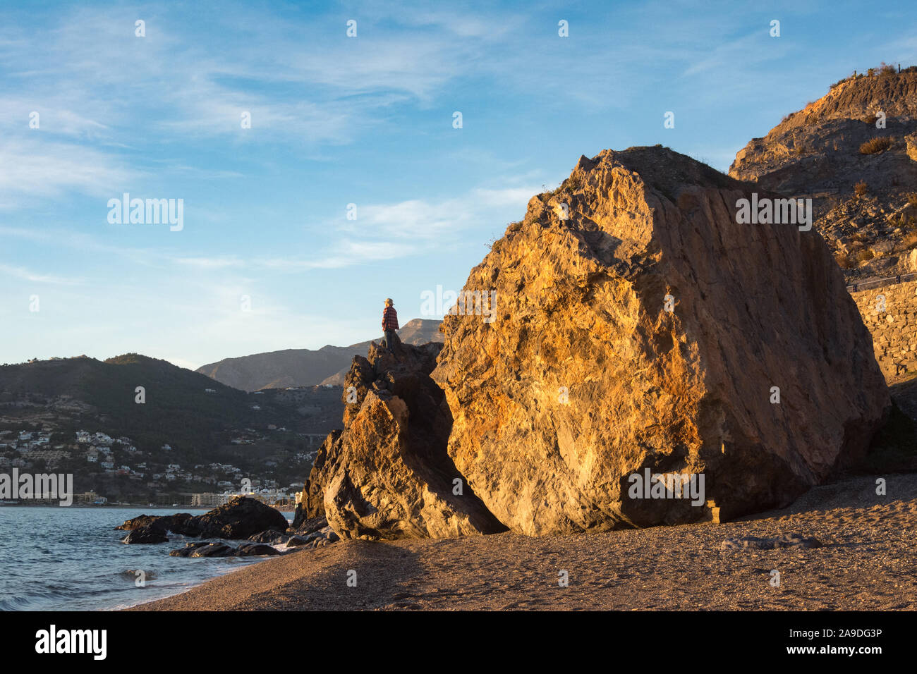 A man stands on a huge boulder he has climbed, looking out to sea on La Herradura beach in Spain. Stock Photo