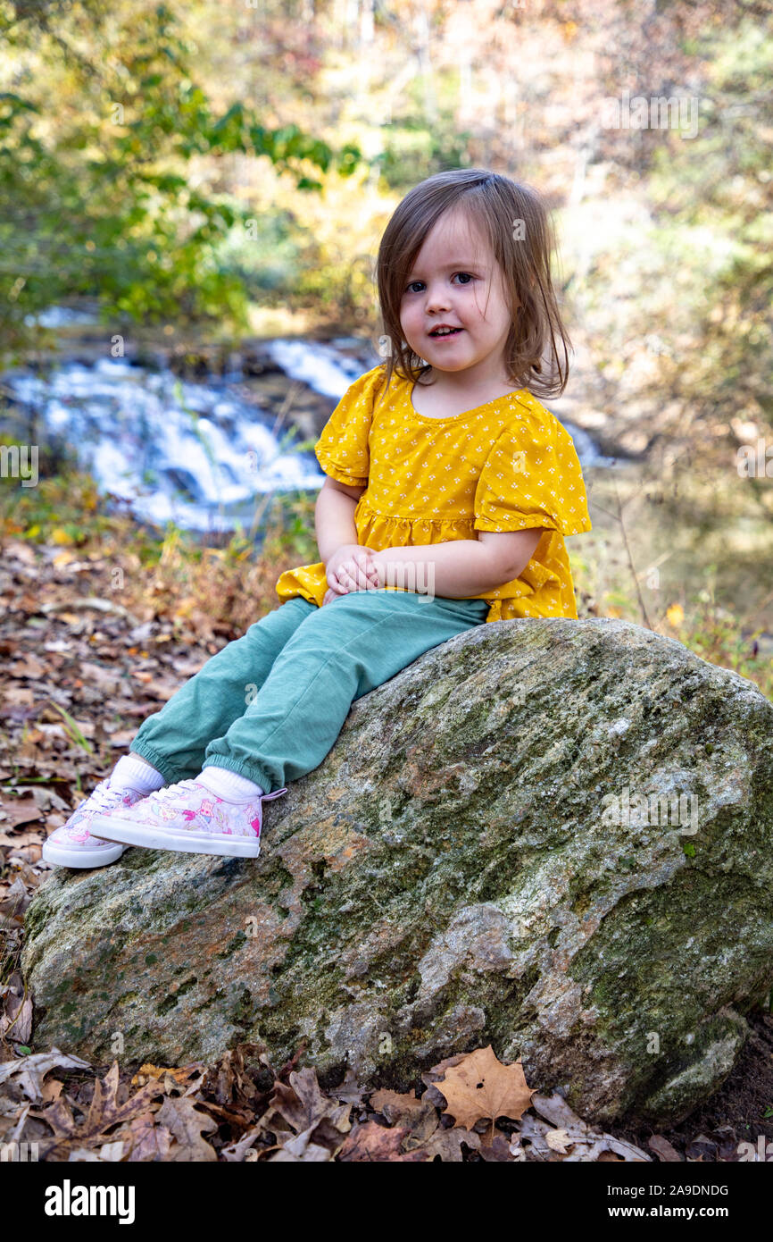 Toddler smiling while sitting on a rock next to waterfall Stock Photo