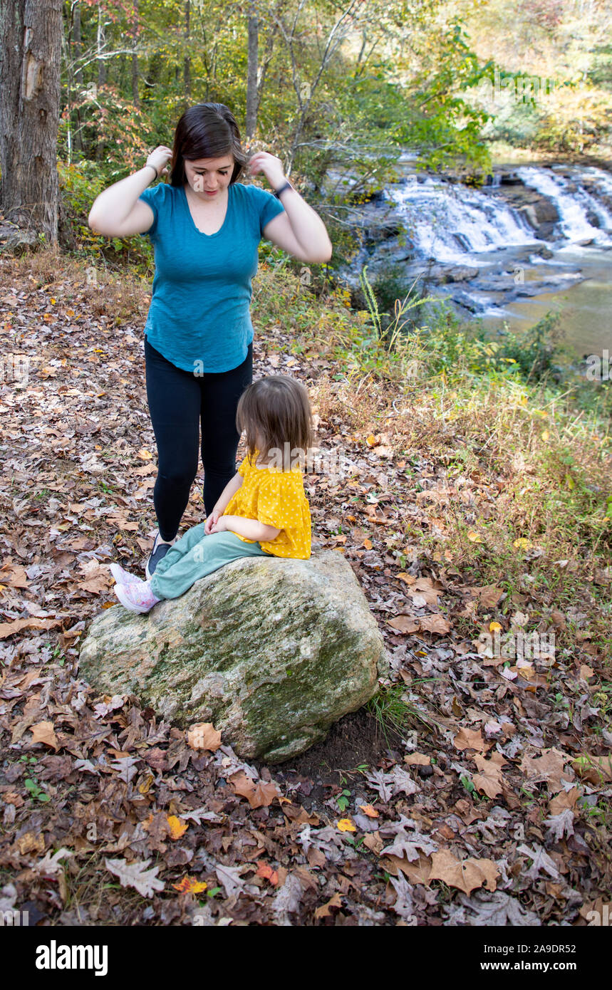 Mother helping daughter while she sits on rock by waterfall Stock Photo