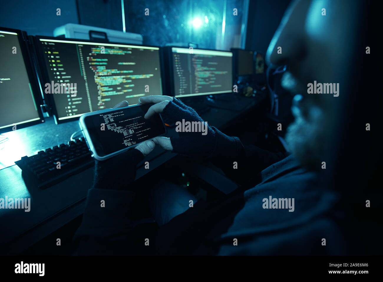 Close-up of computer hacker looking at computer monitor and guessing the password using his mobile phone in dark room Stock Photo